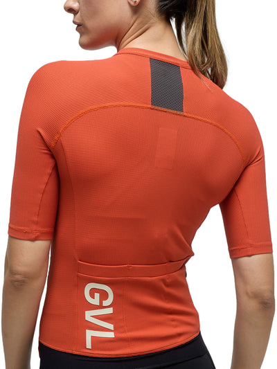 Rear view of a woman in a pumpkin-colored Givelo Modern Classic jersey, illustrating the mesh panel for ventilation, the sturdy zipper design, and the 'GVL' logo on the lower back, ready for high-performance cycling.