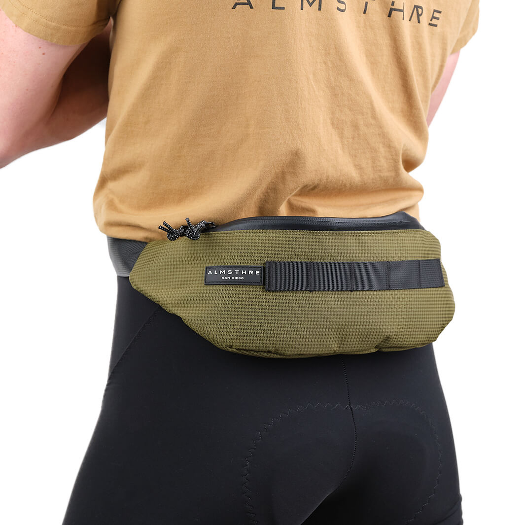Person wearing olive green ALMSTHRE Fanny Pack with black strap and logo tag against white background