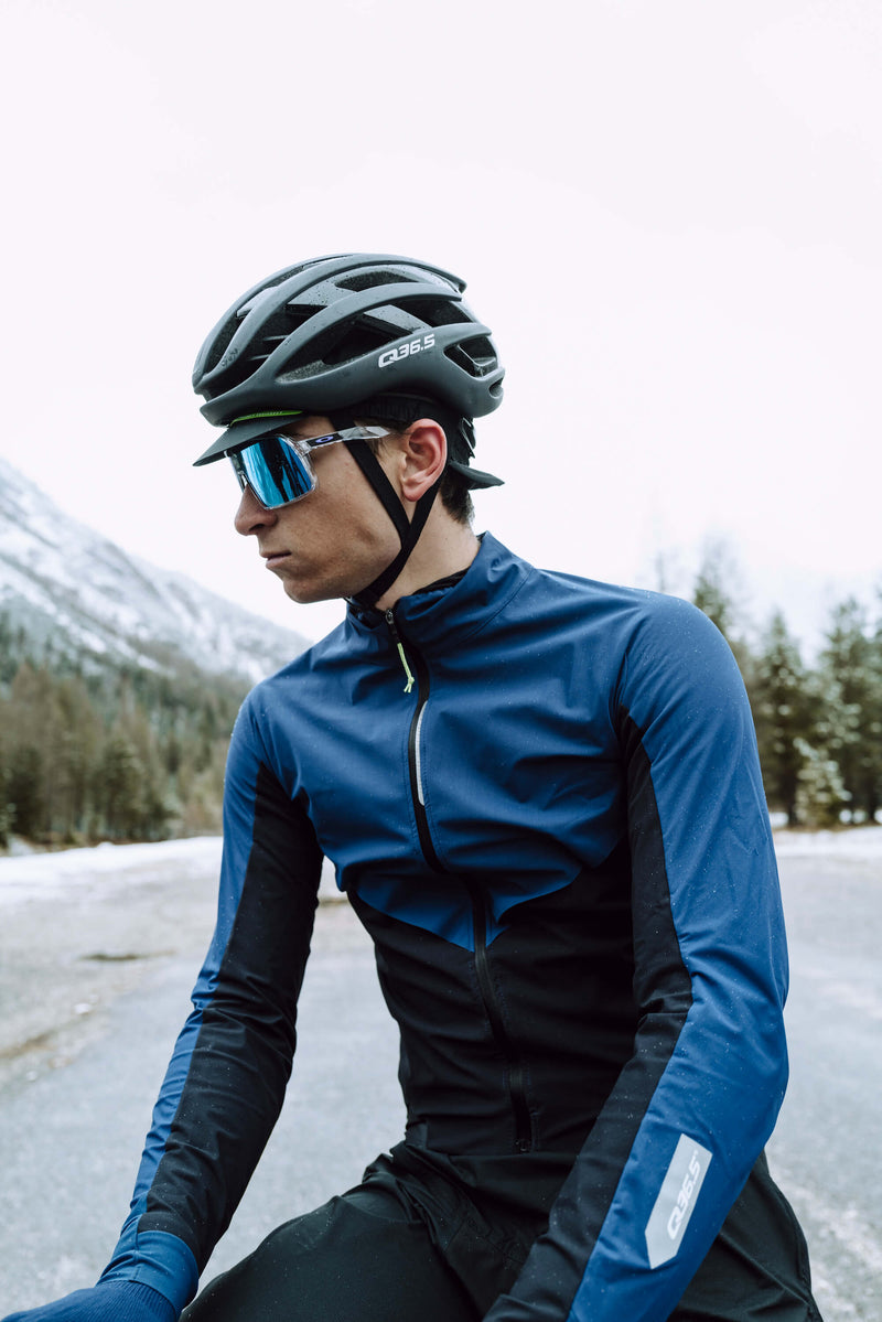 A cyclist wearing the blue Q36.5 Bat Shell Long Sleeve Jersey, demonstrating its fit and temperature control features in a mountainous setting.