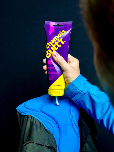 An image of an 8oz bottle of Unisex Chamois cream by Chamois Butt'r being squeezed onto a cycling bib chamois, this is a premium chamois cream for any cyclist.