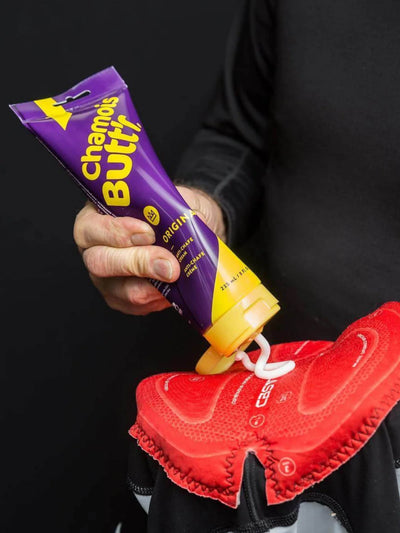 An image of an 8oz bottle of Unisex Chamois cream by Chamois Butt'r being squeezed onto a cycling bib chamois, this is a premium chamois cream for any cyclist.