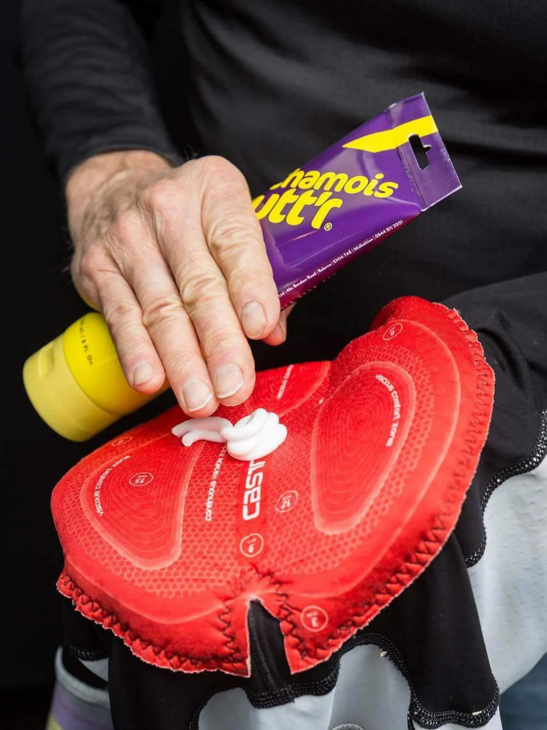 Person applying Chamois Butt'r Chamois Cream - Original onto red padded cycling shorts for comfort and chafe prevention