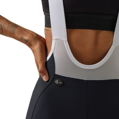 Close-up side view of a woman's lower torso wearing black Givelo HD Pro Bib Shorts, showing the detailed stitching and fabric texture, along with the white shoulder straps and the brand's button logo near the hip.