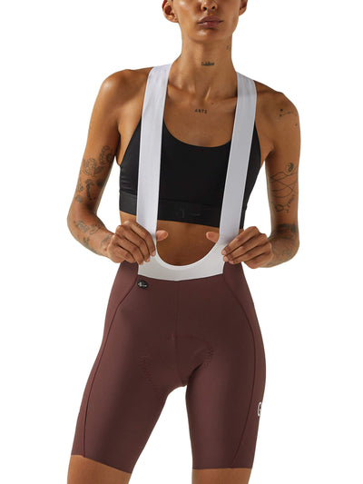 Front view of a woman wearing wine-colored Givelo HD Pro Bib Shorts with a black sports bra and white shoulder straps. The design shows a contoured fit for cycling comfort and performance.