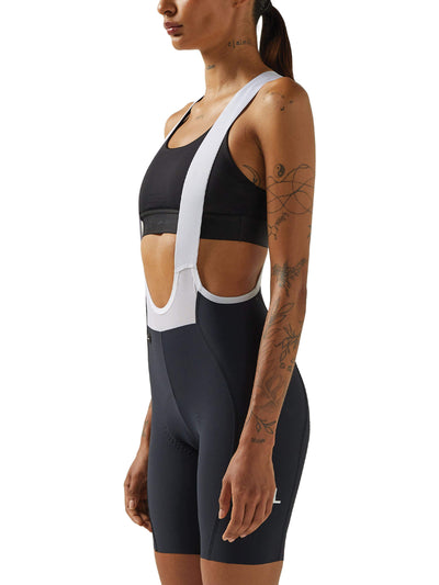 A woman in a side view wearing black Givelo HD Pro Bib Shorts and a white mesh vest. The shorts are designed with attention to comfort for extensive cycling, showcasing the compression and fit that aids in long-duration rides.