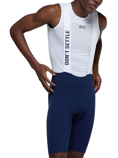 A man posing to the side with his hand on his hip, wearing white Givelo Mayfly bib shorts with a "DON'T SETTLE" slogan, paired with navy blue cycling shorts.
