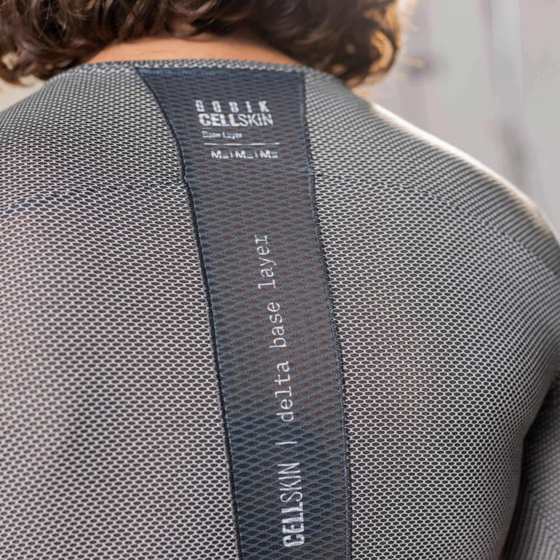 The back view of a man wearing a grey, short-sleeved base layer cycling shirt. A central black stripe with the text 