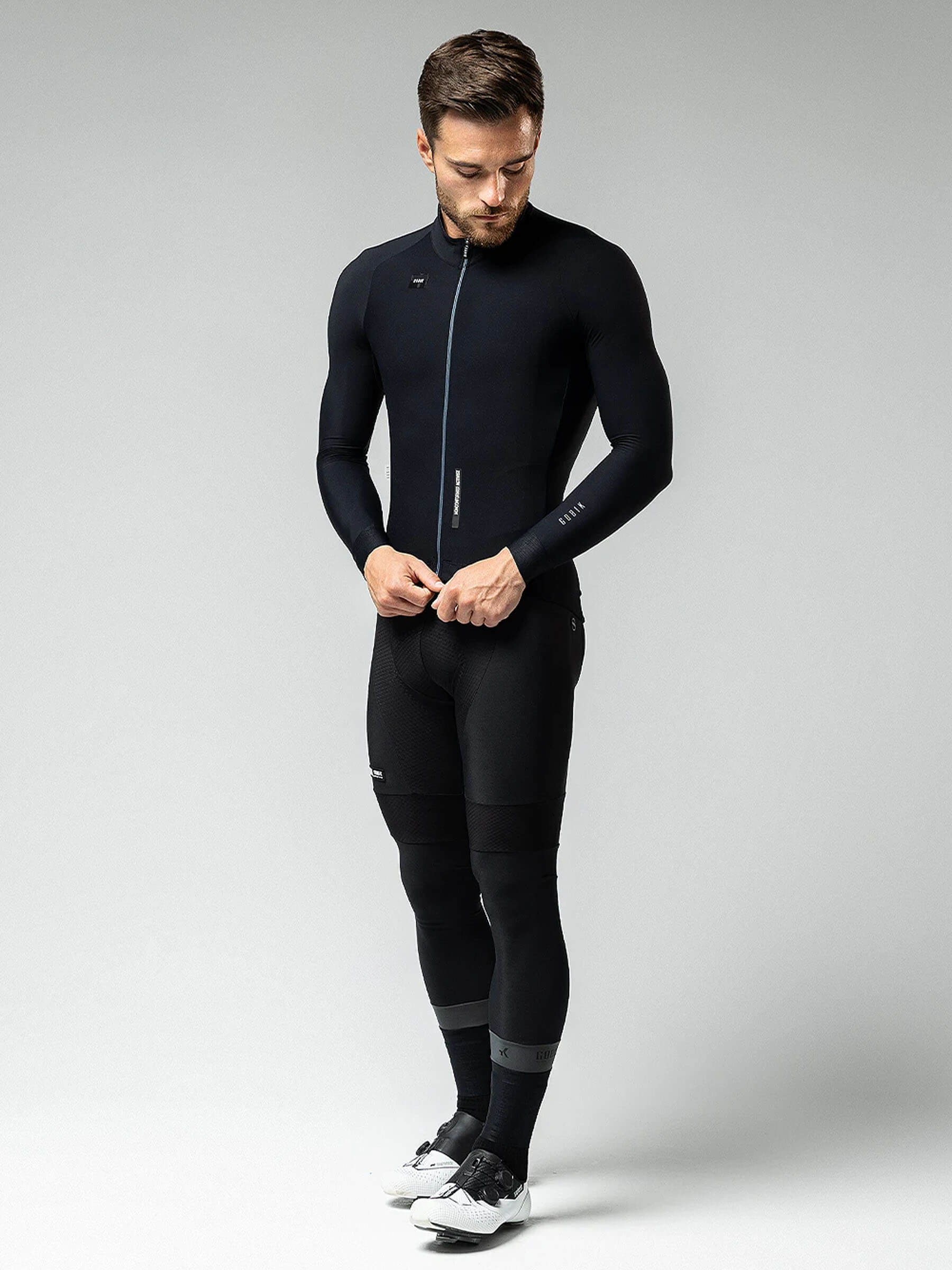 Male cyclist wearing GOBIK Pacer Solid Long Sleeve Jersey - Men's in black, adjusting zipper in minimalist studio setting