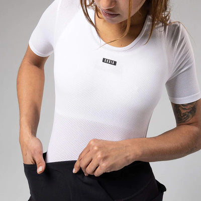 A woman standing in a studio wearing a GOBIK women's short-sleeved base layer in white with a v-neck bib shorts design, ready for a cycling session.