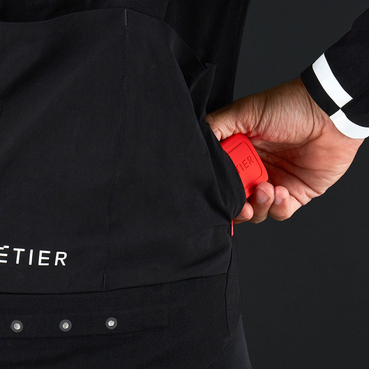 Close-up of a hand placing a red LED wearable device into the back pocket of a black Metier Beacon cycling jacket