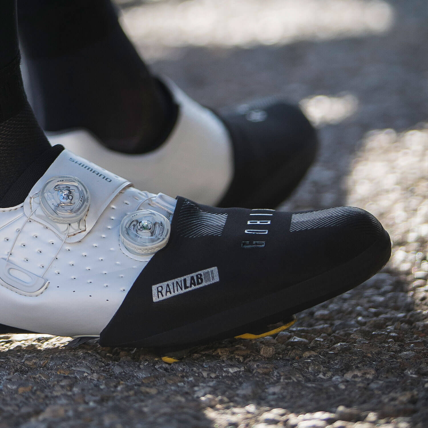 Close-up of a cyclist wearing GOBIK Neoprene Toe Covers Defender Black 2.0 on white cycling shoes on pavement.
