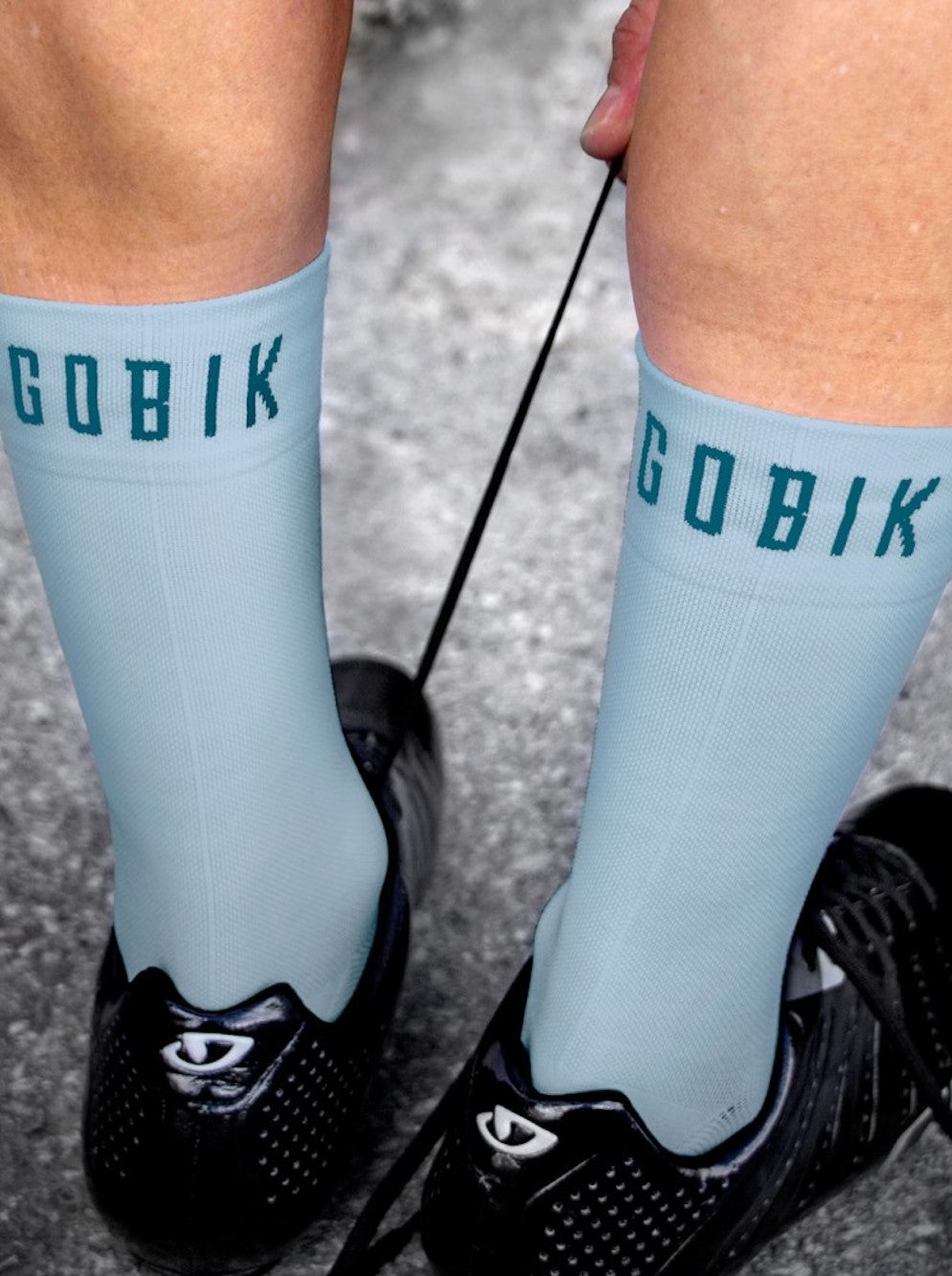 Close-up of feet wearing light blue GOBIK Pure Stone Socks being tied with black cycling shoes on a textured ground